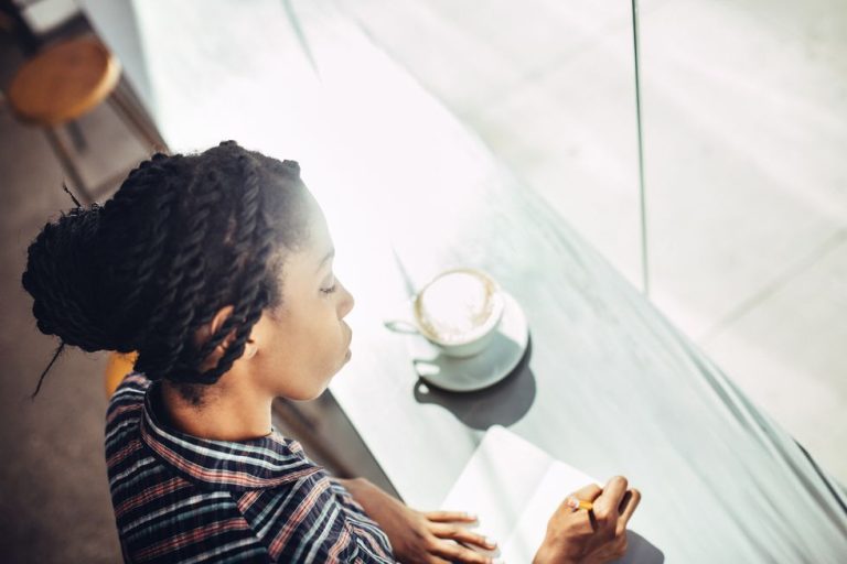 African American Woman Sits At The Window Counter Of A Coffee Shop Enjoying A Latte While Writing Ideas Down In A Small Notepad.jpg