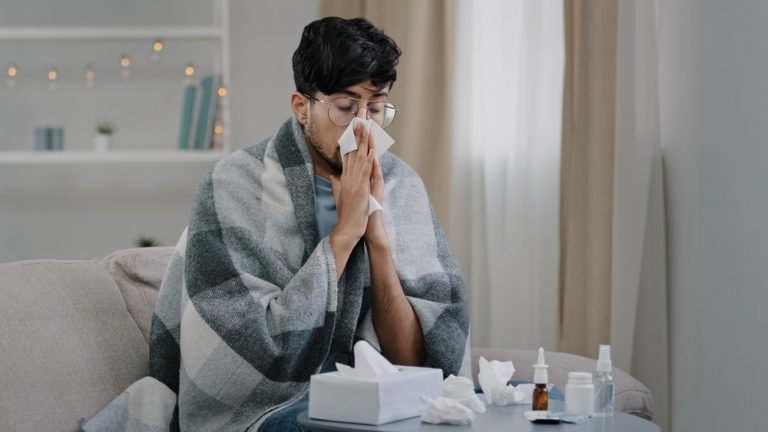 Man On Couch With A Blanket Blowing Nose Has A Cold.jpg