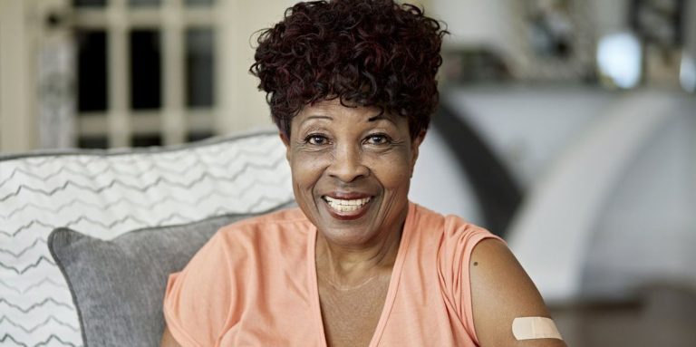 Woman In Casual Clothing With Visible Adhesive Bandage From Vaccination Sitting On Sofa In Family Home And Smiling At Camera.jpg