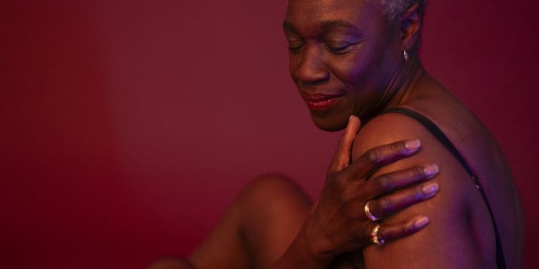 Shorthaired Woman In Semi Dress Seated Against Rose Colored Background Hand On Upper Arm Eyes Closed.jpg