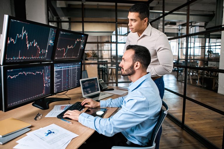 Two Investors Looking At A Series Of Computer Screens With Price Charts On Them.jpg